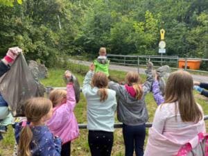 Schulkinder stehen von hinten fotografiert an einem Geländer am Waldrand und halten gefüllte Müllbeutel in die Höhe.